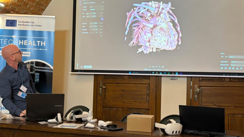 Ekspert prezentuje na ekranie trójwymiarową rekonstrukcję anatomiczną serca i naczyń krwionośnych. Na stole przed nim leżą urządzenia do wirtualnej rzeczywistości i sprzęt komputerowy. W tle baner TECHHEALTH