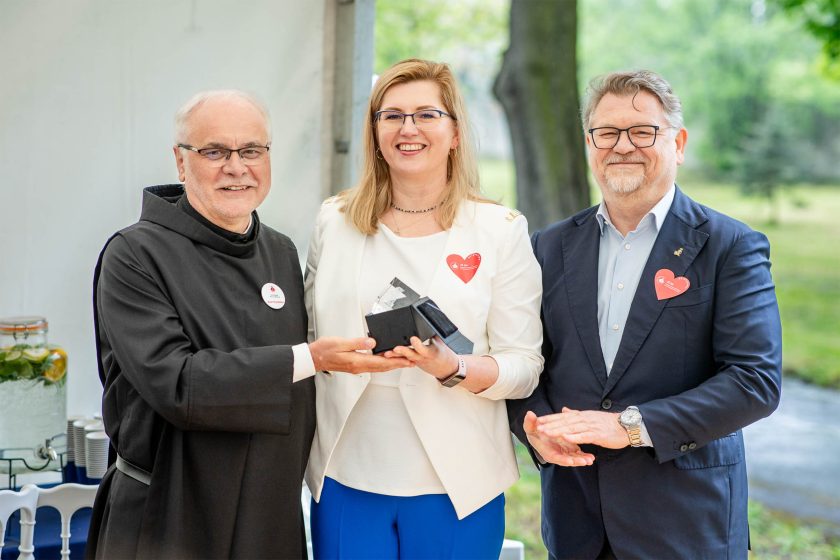 Uśmiechnięta kobieta w białej marynarce odbiera nagrodę – Brat zakonny wręcza nagrodę kobiecie w białej marynarce i niebieskich spodniach. Obok niej stoi mężczyzna w garniturze, wszyscy uśmiechnięci