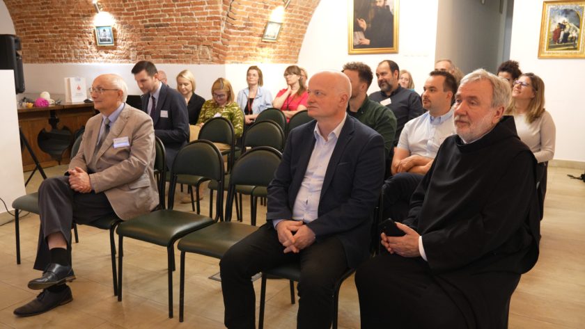 Szerszy kadr na salę konferencyjną. Uczestnicy spotkania siedzą rzędami, w pierwszym rzędzie mężczyźni w garniturach i zakonnik. W tle kolejne rzędy słuchaczy oraz ceglane sklepienia pomieszczenia, ozdobione obrazami i lampami
