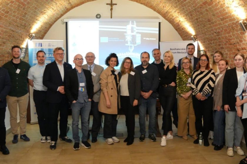 Uczestnicy międzynarodowego projektu Erasmus+ TECHHEALTH zgromadzeni na wspólnym zdjęciu w sali konferencyjnej Bonifraterskiego Centrum Medycznego. Uśmiechnięte twarze, tło stanowią prezentacja multimedialna oraz banery promujące projekt