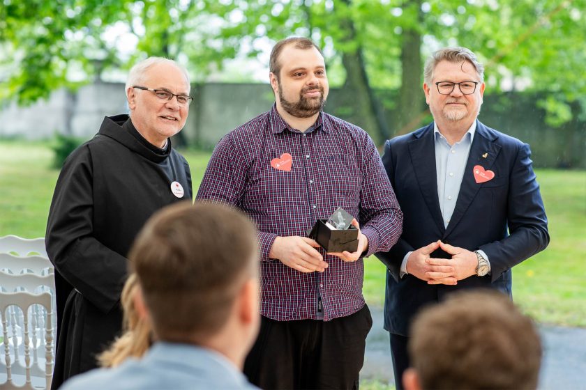 Mężczyzna w kraciaszce z nagrodą – Brat zakonny i mężczyzna w bordowej koszuli w kratę z nagrodą stoją obok siebie, obok nich znajduje się mężczyzna w garniturze. Tło stanowi zielony park