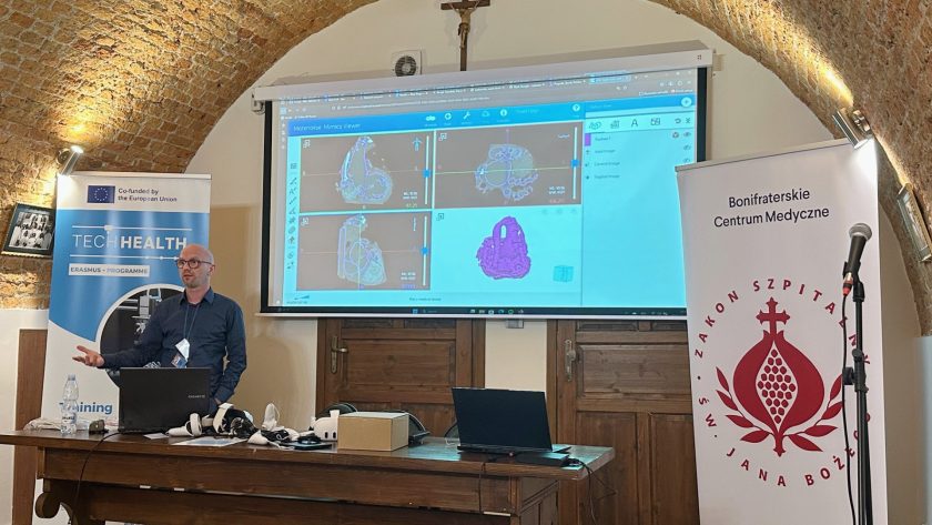 Mężczyzna w ciemnej koszuli i identyfikatorem na szyi prowadzi prezentację w sali wykładowej Bonifraterskiego Centrum Medycznego w ramach projektu Erasmus+ TECHHEALTH. Na ekranie za nim wyświetlony jest interfejs programu do analizy obrazów medycznych – widoczne są trójwymiarowe przekroje czaszki w różnych płaszczyznach. Na stole przed prelegentem leżą gogle rzeczywistości wirtualnej, laptop, akcesoria i woda. Po bokach ekranu znajdują się banery z logotypami projektu oraz Zakonu Szpitalnego św. Jana Bożego. Ceglane sklepienia sali tworzą zabytkową, stylową atmosferę wydarzenia.