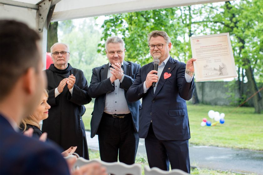 Uhonorowanie na scenie. Mężczyzna w garniturze z mikrofonem w jednej dłoni, drugą unosi w górę dyplom z podziękowaniami. Obok niego dwóch mężczyzn bije brawo – zakonnik i gość w garniturze. W tle zielone drzewa i balony w kolorach biało-czerwono-niebieskich
