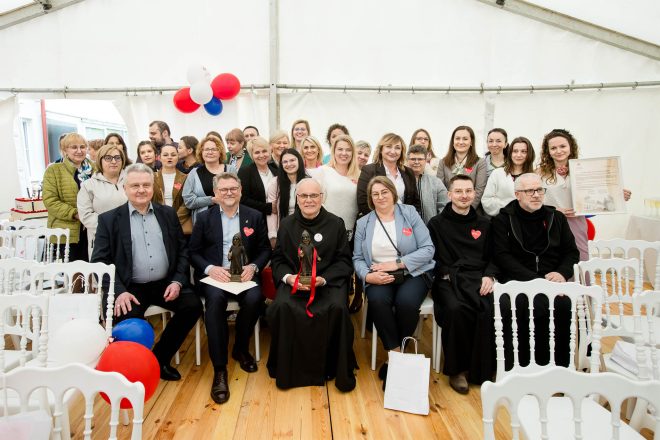 Wspólne zdjęcie uczestników jubileuszu. W centralnej części zdjęcia osoby siedzące w pierwszym rzędzie trzymają statuetki i dyplomy. Za nimi stojący tłum uśmiechniętych pracowników i gości hospicjum. Dekoracja z balonów i tort w tle podkreślają świąteczną atmosferę