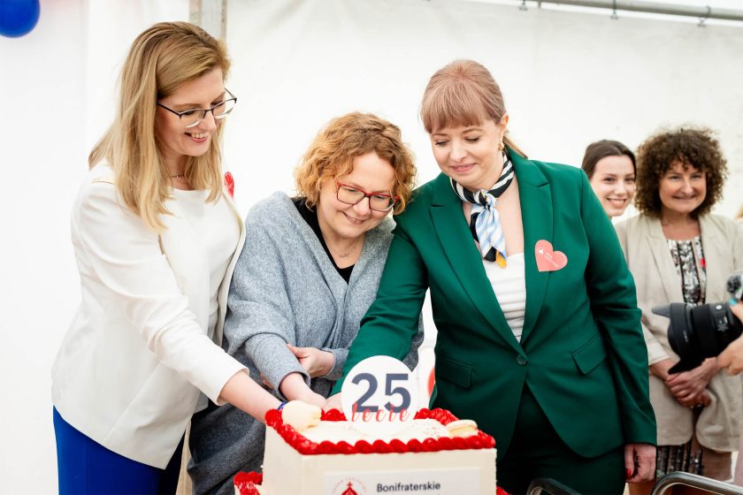 Uroczyste krojenie tortu jubileuszowego. Trzy kobiety wspólnie kroją tort z napisem „25-lecie”. Na ich twarzach widoczna radość i duma. Za nimi grupa gości patrzy z uśmiechem, niektórzy trzymają aparaty