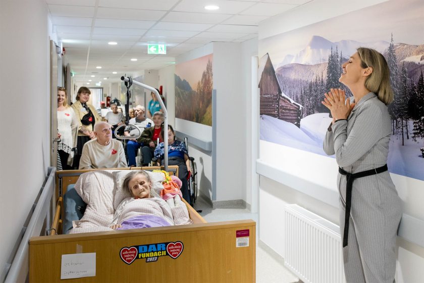 Występ artystyczny na oddziale. Kobieta w szarej sukience z wyrazem wzruszenia na twarzy śpiewa lub recytuje wzdłuż szpitalnego korytarza, w którym ustawieni są pacjenci na wózkach. Na pierwszym planie pacjentka w łóżku, a w tle opiekunowie i pozostali pacjenci