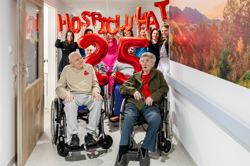 Radosna scena z balonami. Grupa pacjentów i pracowników hospicjum świętuje jubileusz – w tle trzymają czerwone balony układające się w napis „HOSPICJUM 25 LAT”. Na pierwszym planie seniorzy na wózkach patrzą z zaciekawieniem