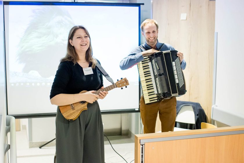 Muzycy przygotowujący się do występu. Kobieta z ukulele i mężczyzna z akordeonem uśmiechają się do publiczności. W tle biały ekran i projektor – miejsce przygotowane do muzykoterapii lub występu