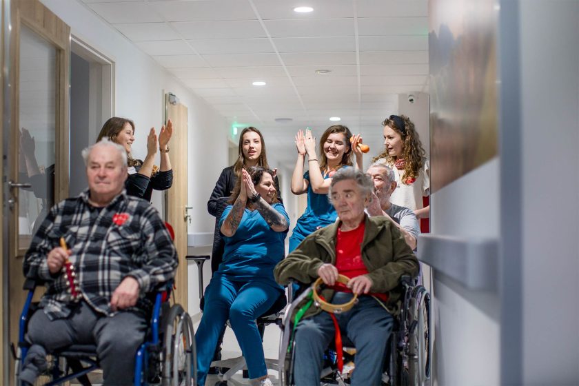 Wspólna zabawa pacjentów i personelu. Na korytarzu hospicjum seniorzy na wózkach grają na instrumentach rytmicznych, a pracownicy i wolontariusze stojący za nimi klaszczą i śpiewają. Panuje radosna, integracyjna atmosfera