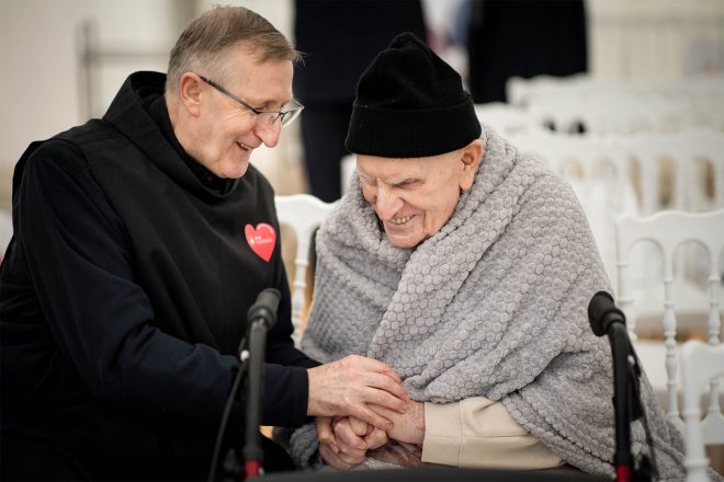 Wzruszający moment czułości. Starszy pacjent otulony szarym kocem uśmiecha się serdecznie, trzymając za rękę zakonnika. Oboje patrzą na siebie z ciepłem i radością, w tle białe krzesła i dekoracje z jubileuszu