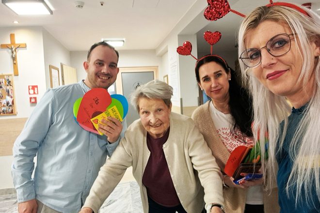 Grupowe zdjęcie wykonane w jasnym korytarzu placówki. W centrum starsza kobieta z siwymi włosami, ubrana w kremowy kardigan, opiera się o chodzik i uśmiecha się do obiektywu. Po jej lewej stronie mężczyzna w jasnoniebieskiej koszuli trzyma w ręku kolorowe serca z napisem „Dla Pani z Oddziału” i „WESOŁYCH WALENTYNEK”. Po prawej stronie stoją dwie kobiety – jedna w czerwonej opasce z brokatowymi serduszkami, druga z jasnymi włosami i okularami, obie trzymają w rękach walentynkowe kartki. W tle widać krzyż na ścianie, tablicę z fotografiami oraz drzwi z napisem sugerującym, że zdjęcie wykonano w przestrzeni wspólnej ośrodka opieki. Ujęcie emanuje życzliwością i międzypokoleniową więzią