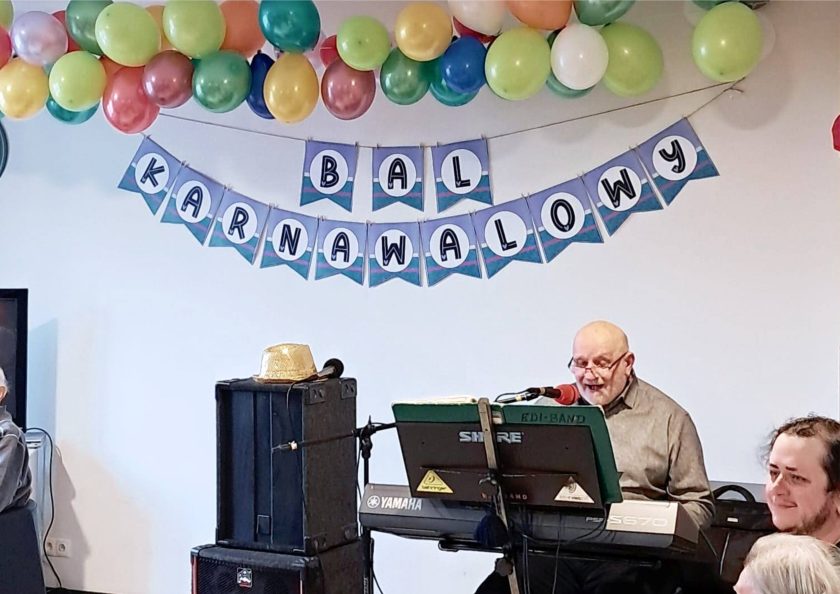 Starszy muzyk grający na keyboardzie podczas balu, uśmiechnięty, śpiewa do mikrofonu. Nad jego głową zawieszona girlanda z balonów i napis „Bal Karnawałowy”. Na głośniku obok keyboardu leży złoty kapelusz. Publiczność widoczna w tle, skupiona na występie