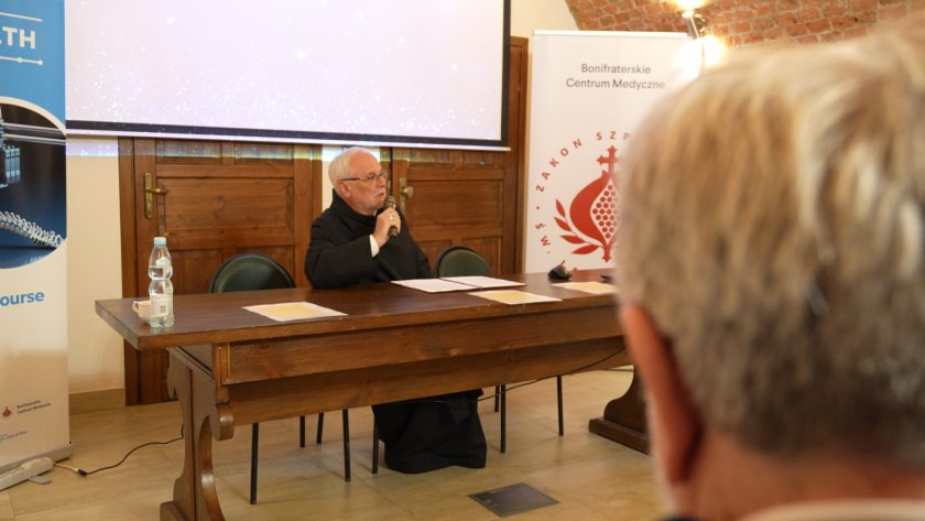 Zakonnik siedzący za stołem prezydialnym przemawia do mikrofonu. Przed nim rozłożone są dokumenty i butelka wody. Po obu stronach stołu stoją roll-upy promujące projekt TECHHEALTH i Bonifraterskie Centrum Medyczne