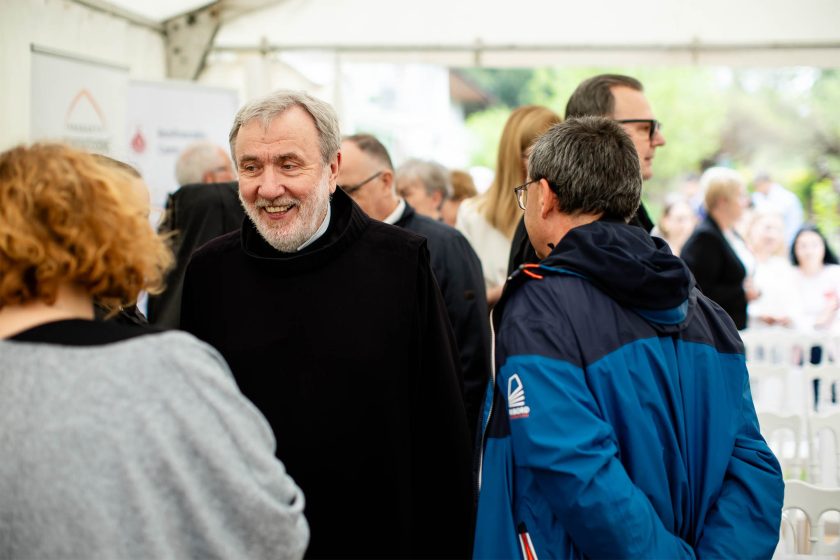 Zadowolony zakonnik w czarnym habicie rozmawia z gośćmi. Scena ma miejsce pod namiotem, wśród wielu uczestników wydarzenia. Na pierwszym planie ujęta jest kobieta z rudymi włosami odwrócona tyłem. Tło rozmyte, ale widać ludzi prowadzących rozmowy i uśmiechających się