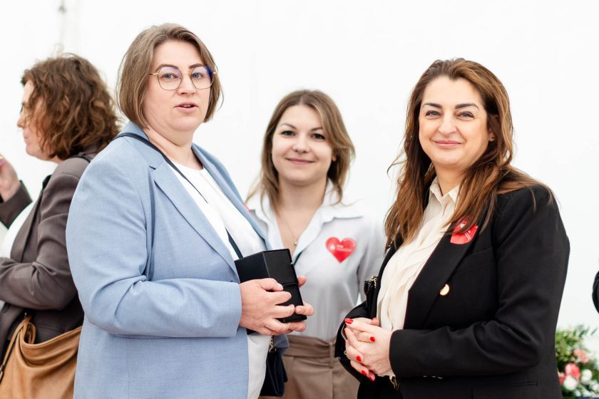 Trzy kobiety stojące razem w rozmowie. Po lewej kobieta w błękitnej marynarce trzyma czarne pudełko z nagrodą, obok niej dwie kobiety w eleganckich strojach z przypiętymi czerwonymi sercami – oznakami wydarzenia jubileuszowego. Białe tło namiotu podkreśla ich sylwetki