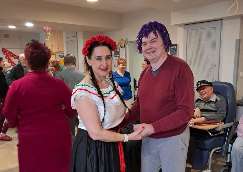 Para taneczna – kobieta w tradycyjnym stroju z czerwonym wiankiem i kolorowymi kolczykami oraz mężczyzna w fioletowej peruce i czerwonym swetrze. Za nimi na fotelu relaksacyjnym siedzi starszy pan w czapce kapitańskiej, uśmiechnięty, obserwujący zabawę. W tle bawiący się uczestnicy i barwne dekoracje ścienne