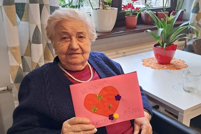 Starsza kobieta z krótkimi, siwymi włosami, ubrana w granatowy sweter i bordową bluzkę z naszyjnikiem z pereł, siedzi przy stole w pobliżu okna i prezentuje własnoręcznie wykonaną walentynkową kartkę. Kartka jest różowa, ozdobiona dużym czerwonym sercem oraz kolorowymi kwiatkami i cekinami, z napisem „WESOŁYCH WALENTYNEK” oraz datą „14.II.2025”. W tle widoczne są zasłony w geometryczne wzory, kilka doniczek z kwiatami oraz szklanka na stole. Seniorka patrzy spokojnie w stronę aparatu, z dumą prezentując efekt swojej pracy