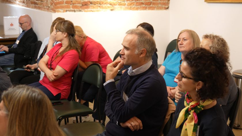 Uczestnicy spotkania siedzący w rzędach. Na pierwszym planie mężczyzna z siwiejącymi włosami, w granatowym swetrze, trzymający rękę przy brodzie w zamyślonej pozie. Obok kobieta z kolorową apaszką. W tle kobiety w ubraniach medycznych i codziennych