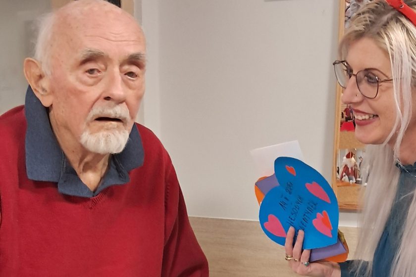 Starszy mężczyzna z siwą brodą i wąsami, ubrany w bordowy sweter z kołnierzem w odcieniu granatu, siedzi przy stole i patrzy w kierunku aparatu. Obok niego, uśmiechnięta kobieta z długimi blond włosami, w okularach i z opaską z czerwonymi sercami, pokazuje mu kolorową kartkę w kształcie niebieskiego serca z napisem „14.II.2025 WESOŁYCH WALENTYNEK”, ozdobioną czerwonymi serduszkami. Atmosfera jest ciepła i serdeczna, a kadr uchwyca moment radosnej wymiany walentynkowych życzeń