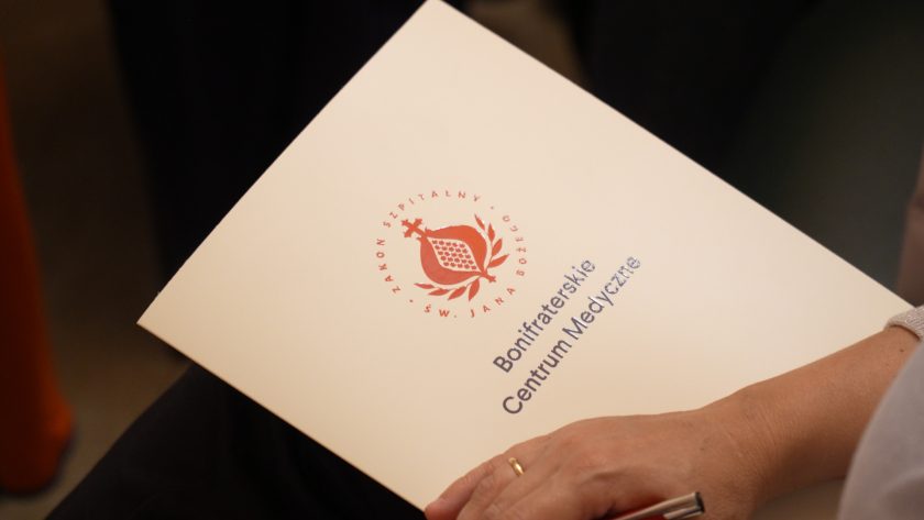Zbliżenie na dłoń osoby trzymającej teczkę z logo Bonifraterskiego Centrum Medycznego oraz charakterystycznym granatowo-czerwonym znakiem Zakonu Szpitalnego św. Jana Bożego. W tle ciemne tło i fragment odzieży uczestnika