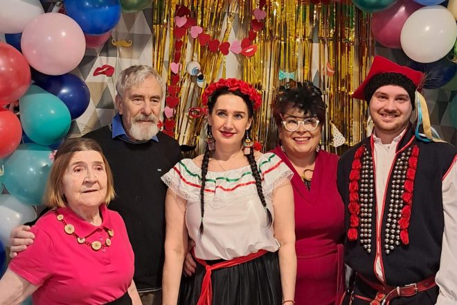 Pamiątkowe zdjęcie grupowe uczestników balu karnawałowego – na tle kolorowych balonów i złotej, błyszczącej dekoracji z elementami masek i ust. W centrum kobieta w meksykańskim stroju, obok niej seniorka w różowej koszulce, mężczyzna z siwą brodą, energiczna prowadząca w czerwonej sukience oraz mężczyzna w stroju ludowym. Wszyscy uśmiechnięci, pozujący do wspólnej fotografii