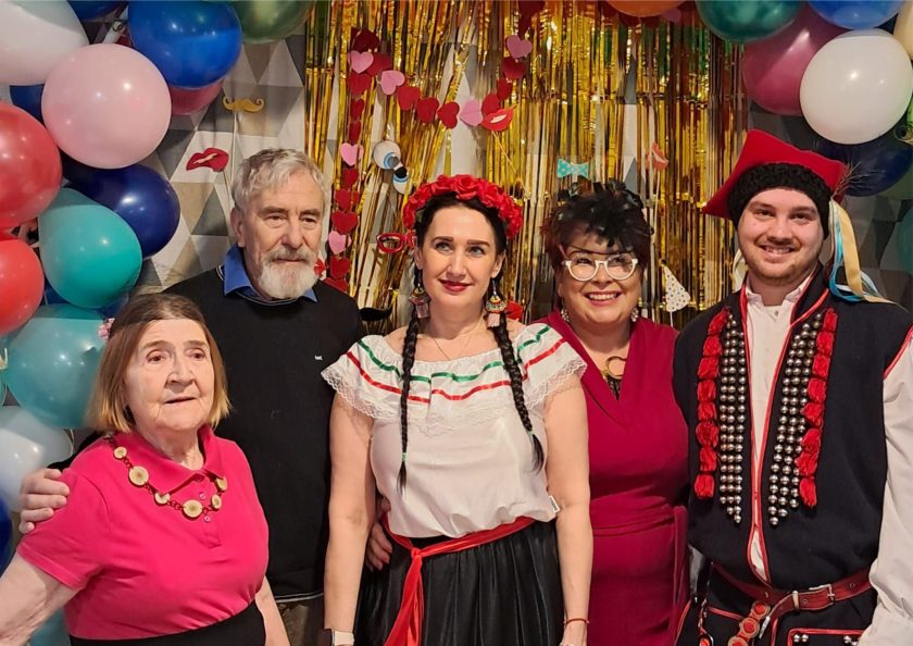 Pamiątkowe zdjęcie grupowe uczestników balu karnawałowego – na tle kolorowych balonów i złotej, błyszczącej dekoracji z elementami masek i ust. W centrum kobieta w meksykańskim stroju, obok niej seniorka w różowej koszulce, mężczyzna z siwą brodą, energiczna prowadząca w czerwonej sukience oraz mężczyzna w stroju ludowym. Wszyscy uśmiechnięci, pozujący do wspólnej fotografii