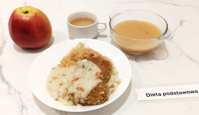 Zestaw obiadowy oznaczony jako „Dieta podstawowa”, podany na białym blacie. Na pierwszym planie biały talerz z porcją ziemniaków puree, surówką z kiszonej kapusty i panierowanym kotletem schabowym oblanym białym sosem z warzywami. W tle znajduje się miseczka z jasną zupą jarzynową o półpłynnej konsystencji, filiżanka z herbatą z mlekiem oraz czerwone jabłko jako dodatek owocowy. Całość prezentuje się jako pełnowartościowy, klasyczny polski obiad
