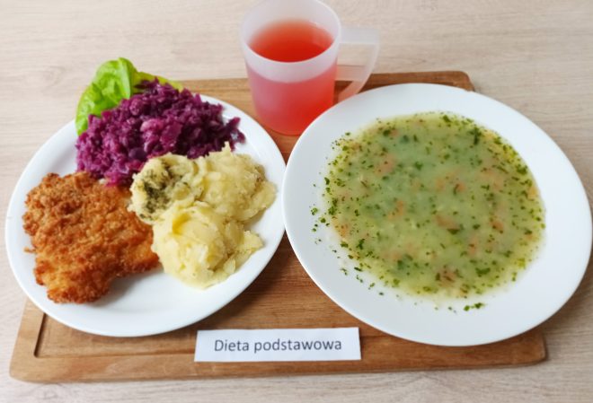 Na drewnianej tacy podano klasyczny obiad. Na jednym talerzu – panierowany kotlet schabowy, ziemniaki puree z koperkiem i czerwona kapusta zasmażana, udekorowana liściem sałaty. Na drugim talerzu – zupa jarzynowa z ziemniakami i natką pietruszki. Obok przezroczysty kubek z czerwonym napojem. Całość podpisana jako „Dieta podstawowa”