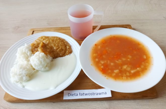 Zestaw obiadowy w diecie łatwostrawnej podany na białych talerzach na drewnianej tacy. Danie główne składa się z dwóch kopczyków ryżu, polanych białym sosem jogurtowym oraz porcji rozdrobnionej pieczeni lub pulpetu w jasnym sosie. Obok miska z zupą jarzynową na klarownym wywarze – widoczne kawałki marchwi, ziemniaków i pietruszki. Do picia różowy napój w przezroczystym kubku z uchem. Posiłek lekkostrawny, bez przypraw drażniących