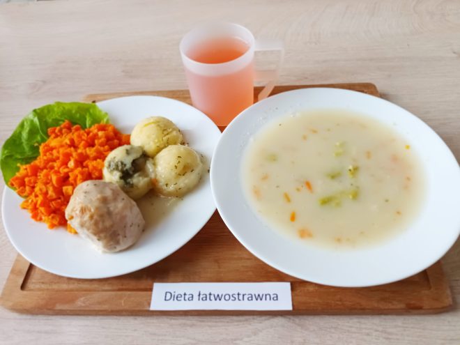 Obiad łatwostrawny złożony z zupy jarzynowej na mlecznej bazie z kawałkami marchwi i brokułów oraz drugiego dania na talerzu: ugotowana marchewka w kostkę, trzy kluski śląskie z sosem ziołowym oraz gotowany pulpet drobiowy. Do picia różowy napój w plastikowym kubku. Całość estetyczna i przygotowana z myślą o osobach wymagających delikatnej diety