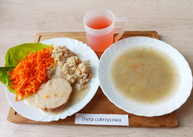 Na jasnobrązowej tacy znajduje się zestaw obiadowy w diecie cukrzycowej. W skład dania głównego podanego na białym talerzu wchodzą: plaster gotowanego mięsa polanego jasnym sosem, porcja kaszy oraz starta marchewka ułożona na liściu sałaty. Obok znajduje się głęboki talerz z klarowną zupą jarzynową, w której widoczne są drobne cząstki warzyw. Posiłek uzupełnia różowy kompot w przezroczystym kubku z uchwytem. Całość ustawiona jest na tacy z podpisem „Dieta cukrzycowa”