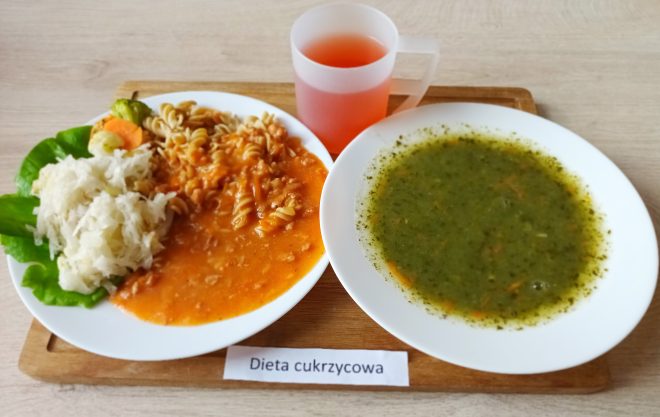Na zdjęciu widoczny jest zestaw obiadowy serwowany na jasnym blacie, ustawiony na drewnianej tacy. Po lewej stronie znajduje się talerz z porcją gotowanego makaronu świderki w czerwonym sosie pomidorowym oraz dużą porcją białej kapusty kiszonej z dodatkiem marchewki i listka sałaty. Obok, po prawej stronie, podano zupę o ciemnozielonym zabarwieniu, najpewniej zupę jarzynową na bazie brokułów lub szpinaku, z dodatkiem przypraw ziołowych. Do obiadu dołączono napój o czerwonym kolorze w przezroczystym kubku z uchem. Całość oznaczona etykietą „Dieta cukrzycowa”, co sugeruje posiłek dostosowany do potrzeb osób z cukrzycą, charakteryzujący się ograniczoną zawartością cukrów prostych