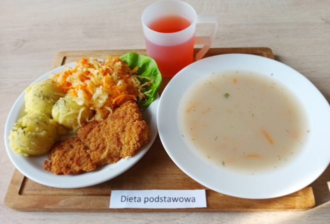 Na tym samym rodzaju tacy umieszczono posiłek oznaczony etykietą „Dieta podstawowa”. Zestaw obiadowy składa się z dwóch białych talerzy. Na pierwszym, większym, znajdują się trzy porcje ziemniaków z koperkiem, panierowany i smażony filet z mięsa (prawdopodobnie schabowy) oraz mieszanka surówek z kapusty i marchwi, podana na liściu sałaty. Obok, na mniejszym talerzu, widoczna jest miska z jasną, lekko zawiesistą zupą jarzynową z widocznymi fragmentami marchwi i ziół. Do zestawu dołączono także przezroczysty kubek z czerwonym napojem, przypominającym kompot owocowy. Posiłek prezentuje się jako pełnowartościowy, klasyczny obiad
