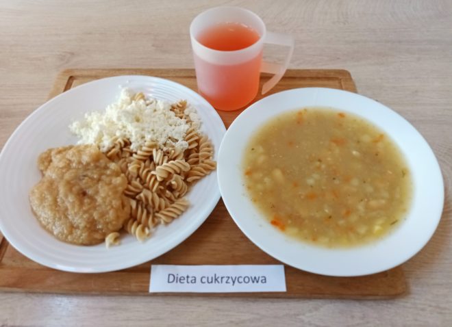 Taca z obiadem w diecie cukrzycowej. Na białym talerzu znajduje się porcja pełnoziarnistego makaronu świderki, posypanego białym, rozdrobnionym twarogiem oraz kleik z jabłek lub duszonych owoców. Obok stoi talerz z gęstą zupą jarzynową – widoczne są w niej drobno pokrojone warzywa: marchew, ziemniaki i przyprawy. Do posiłku podano czerwonawy napój w przezroczystym kubku z uchwytem. Całość ustawiona na drewnianej tacy z białą etykietą „Dieta cukrzycowa”
