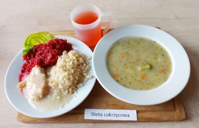 Taca z obiadem dostosowanym do potrzeb osób z cukrzycą, ustawiona na jasnym blacie. Po lewej stronie znajduje się duży płaski talerz, na którym podano porcję gotowanej kaszy jęczmiennej, filet z kurczaka w jasnym sosie oraz buraczki na ciepło – drobno starte i ułożone na liściu sałaty. Obok kaszy widoczna jest niewielka ilość sosu z mięsa. Po prawej stronie ustawiono głęboki talerz z zupą jarzynową – w lekkim, jasnozielonym wywarze unoszą się kawałki marchewki, ziemniaków i brokułów. W tle stoi półprzezroczysty kubek z różowym kompotem. Na tacy umieszczono również etykietę z napisem „Dieta cukrzycowa”