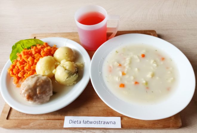 Na zdjęciu widoczny jest posiłek obiadowy przeznaczony dla osoby na diecie łatwostrawnej, podany na jasnej tacy z drewnianą podstawką i podpisem „Dieta łatwostrawna”. Po lewej stronie znajduje się talerz z gotowaną marchewką pokrojoną w drobną kostkę, trzema ziemniakami posypanymi koperkiem oraz dużym klopsikiem z mięsa mielonego, polanym delikatnym sosem. Całość uzupełnia zielony liść sałaty. Po prawej stronie tacy widnieje talerz z jasną, lekko zawiesistą zupą warzywną z makaronem świderki i drobną kostką marchewki oraz pietruszki. W tle stoi przezroczysty kubek z czerwonym napojem owocowym. Estetycznie podany posiłek sugeruje dietę lekkostrawną, odpowiednią dla pacjentów wymagających delikatnego odżywiania
