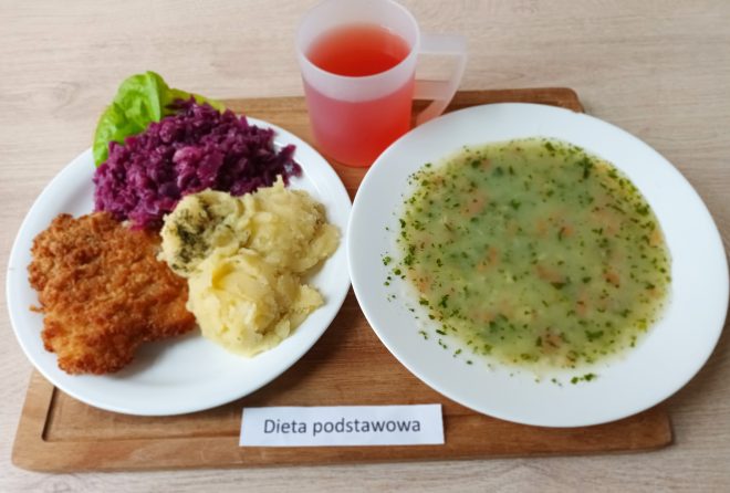 Na drewnianej tacy ułożono szpitalny zestaw obiadowy oznaczony jako „Dieta podstawowa”. Na dużym talerzu po lewej stronie znajduje się panierowany kotlet schabowy, porcja tłuczonych ziemniaków posypanych koperkiem oraz gotowana czerwona kapusta. Danie ozdobiono liściem sałaty. Obok, na drugim talerzu, podano zupę jarzynową o gęstej konsystencji, z widocznymi cząstkami marchewki, ziemniaków i zieleniny. Do obiadu dołączono przezroczysty kubek z czerwonym kompotem lub herbatą owocową