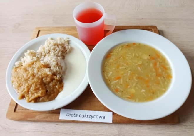 Taca z posiłkiem oznaczonym jako dieta cukrzycowa, ustawiona na tym samym jasnym blacie. Na tacy znajdują się: Talerz obiadowy – Zawiera dwie porcje: po jednej stronie ugotowany brązowy ryż oblany musem owocowym (prawdopodobnie jabłkowym) o lekko brązowym zabarwieniu, po drugiej stronie jasny sos mleczny lub jogurt naturalny. Zupa – W osobnym, głębokim talerzu znajduje się jarzynowa zupa z cienkim makaronem. W rosole pływają cienkie paski marchewki, ziemniaka i przyprawy – całość ma złocisty kolor. Napój – Czerwony, przezroczysty płyn (najpewniej napój owocowy bez cukru lub słaba herbata z hibiskusa), podany w matowym plastikowym kubku z uchem. Na środku tacy widoczna jest etykieta z napisem "Dieta cukrzycowa".
