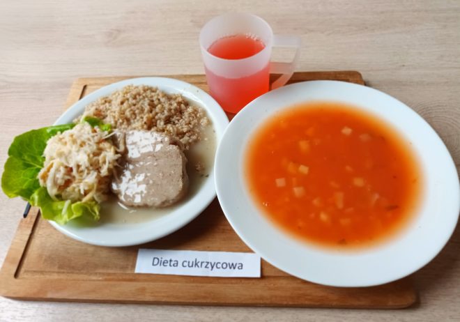 Na drewnianej tacy znajduje się pełnowartościowy obiad przeznaczony dla osoby z cukrzycą. Na głównym talerzu ułożone są trzy składniki: porcja kaszy gryczanej, plaster gotowanego mięsa (najprawdopodobniej wieprzowego lub cielęcego) w jasnym sosie oraz surówka z białej kapusty na liściu sałaty. Obok talerza ustawiono głęboki talerz z czerwoną zupą jarzynową – z widocznymi w niej kawałkami marchwi, ziemniaków i przypraw. Całość uzupełnia przezroczysty, matowy kubek z różowym napojem, najpewniej kompotem bez dodatku cukru. Poniżej znajduje się etykieta z napisem „Dieta cukrzycowa”