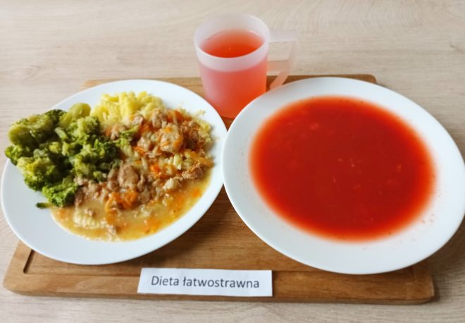 Taca z posiłkiem obiadowym oznaczonym etykietą „Dieta łatwostrawna”. Na niej znajdują się trzy elementy posiłku: na głębokim talerzu po prawej stronie podano klarowną zupę pomidorową o intensywnie czerwonym kolorze, bez widocznych dodatków stałych. Po lewej stronie na płaskim talerzu umieszczono gotowanego brokuła, ziemniaki tłuczone z masłem oraz duszone mięso w lekkim sosie z marchewką i cebulą – danie wygląda na lekkostrawne, przygotowane bez smażenia. Do posiłku dołączono także przezroczysty kubek z czerwonym kompotem. Wszystko ułożone jest na drewnianej tacy stojącej na jasnym blacie