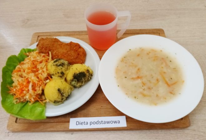 Na drewnianej tacce ułożone są dwa talerze i kubek z różowym napojem. Na głównym talerzu po lewej stronie znajduje się klasyczny zestaw obiadowy: panierowany kotlet schabowy, trzy ziemniaki z dodatkiem koperku oraz surówka z marchwi i białej kapusty, podane na liściu sałaty. Obok, w białej misce, znajduje się zupa z pływającymi w niej warzywami (marchew, ziemniaki, zioła – prawdopodobnie zupa jarzynowa). Wszystko stoi na jasnym blacie, a pod tacką znajduje się podpis „Dieta podstawowa"