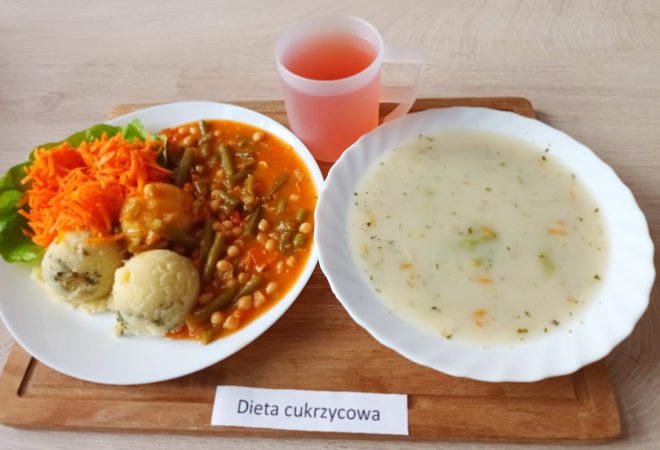 Tacka z dwoma talerzami i różowym napojem w półprzezroczystym kubku. Na głównym talerzu podano gulasz warzywno-strączkowy z zieloną fasolką szparagową i ciecierzycą w sosie pomidorowym, porcję gotowanych ziemniaków z koperkiem oraz startą marchewkę jako surówkę. Wszystko ułożone na liściu sałaty. Obok w talerzu znajduje się zupa krem (najprawdopodobniej brokułowa z dodatkiem warzyw). Danie przeznaczone dla pacjentów z cukrzycą, co potwierdza podpis „Dieta cukrzycowa” umieszczony pod tacką