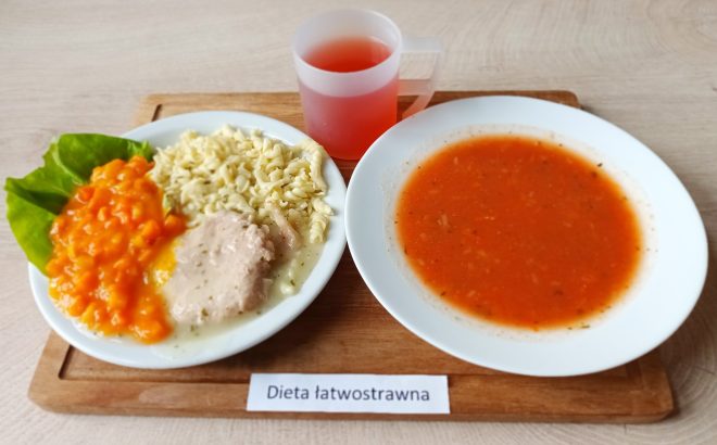 Na jasnej tacy umieszczono dwa talerze i kubek z napojem owocowym. Na większym talerzu makaron świderki z gotowaną cielęciną w jasnym sosie i gotowaną, rozdrobnioną marchewką. Całość uzupełnia liść sałaty masłowej. Na drugim talerzu delikatna, klarowna zupa pomidorowa. Na etykiecie przed tacą widnieje napis „Dieta łatwostrawna”. Posiłek dostosowany do potrzeb osób z zaburzeniami trawienia