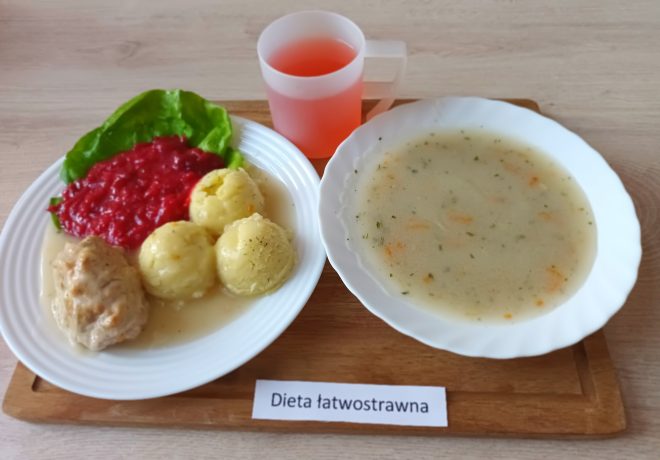 Obiad dla osoby na diecie łatwostrawnej. Na dużym białym talerzu znajdują się trzy kluski ziemniaczane polane jasnym sosem, porcja gotowanego mięsa (najpewniej pulpeta drobiowego) oraz ćwikła z buraków na liściu sałaty. Obok – biały talerz z klarowną zupą warzywną na bazie ziemniaka, marchwi i natki pietruszki. Do picia podano jasnoróżowy napar w półprzezroczystym kubku. Całość zestawu ustawiono na drewnianej tacy z etykietą „Dieta łatwostrawna”