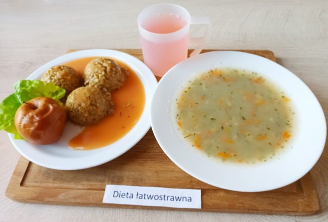 Zestaw obiadowy na jasnej tacy. Z lewej strony talerz z trzema kulkami kaszy gryczanej polanymi łagodnym sosem, dekorowany liściem sałaty oraz pieczonym jabłkiem. Po prawej stronie głęboki talerz z delikatną zupą jarzynową na bazie klarownego bulionu z drobnymi warzywami. Do zestawu dołączony przezroczysty plastikowy kubek z różowym napojem. Etykieta pod spodem głosi: „Dieta łatwostrawna”