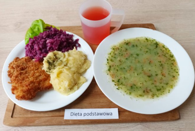 Na drewnianej tacy podano zestaw obiadowy z etykietą "Dieta podstawowa". Po lewej stronie znajduje się talerz z trzema głównymi składnikami: złocistym, panierowanym kotletem schabowym, porcją tłuczonych ziemniaków z odrobiną zieleniny oraz duszoną czerwoną kapustą, udekorowaną liściem sałaty. Po prawej stronie, na osobnym talerzu, znajduje się zupa jarzynowa – klarowna, z drobno pokrojonymi warzywami (m.in. marchewką, ziemniakami i natką pietruszki). W tle stoi półprzezroczysty kubek z czerwonym kompotem. Całość serwisu sprawia wrażenie domowego, zbilansowanego posiłku obiadowego