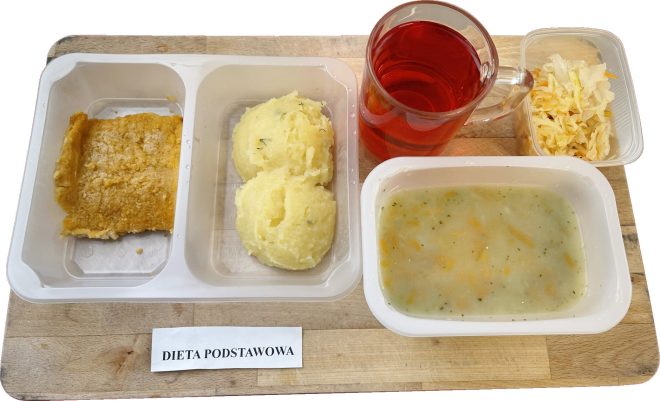 Dieta obiadowa serwowana w białym plastikowym pojemniku. W jednym przedziale znajduje się zapiekanka ziemniaczana lub ryżowa, w drugim – dwa kopce ziemniaków puree z koperkiem. W mniejszym pojemniku – zupa jarzynowa. Dodatkowo: surówka z kapusty i kubek czerwonego napoju. Etykieta informuje: „Dieta podstawowa”