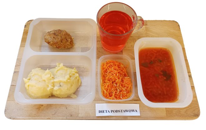 Obiad w ramach diety podstawowej, tym razem na plastikowej tacy. W zestawie: pieczony mielony kotlet, ziemniaki puree z koperkiem, surówka z marchwi oraz zupa jarzynowa z makaronem w oddzielnym pojemniku. Do picia – herbata lub kompot w szklance o czerwonym zabarwieniu. Kolorystyka i skład posiłku sugerują ciepły, domowy charakter, typowy dla szpitalnych obiadów