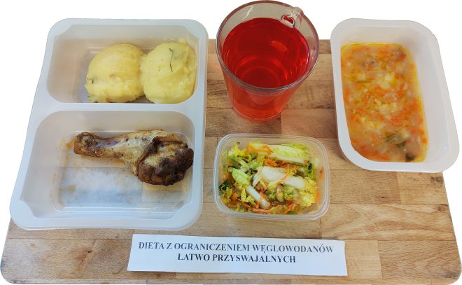 Na drewnianej tacy ułożony jest posiłek składający się z trzech białych pojemników i jednego kubka. W lewym pojemniku znajdują się dwie porcje ziemniaczanego purée z koperkiem oraz pieczone udko kurczaka. Obok, w przezroczystym pudełku, podano surówkę z białej kapusty z dodatkiem marchewki. W prawym górnym rogu znajduje się kubek z czerwonym napojem, a tuż obok – zupa jarzynowa z widocznymi kawałkami marchwi, ziemniaków i zieleniny. Całość opatrzona jest etykietą z napisem: „Dieta z ograniczeniem węglowodanów łatwo przyswajalnych”
