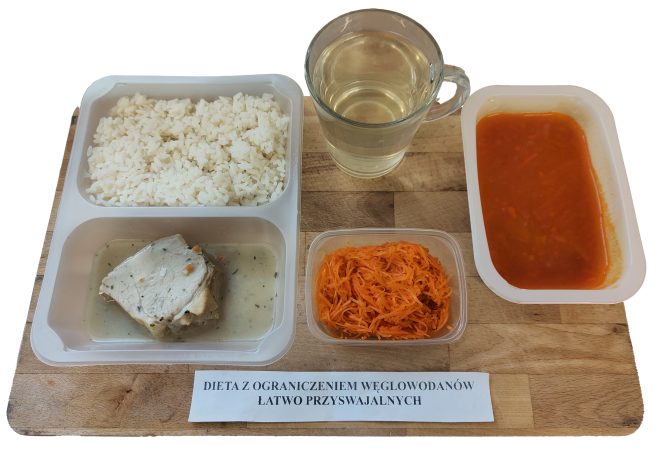Obiad skomponowany z myślą o ograniczeniu szybko przyswajalnych cukrów. W plastikowej tacce znajduje się porcja białego ryżu, kawałek gotowanego mięsa w delikatnym, jasnym sosie oraz surówka z tartej marchewki. Obok umieszczono przezroczysty kubek z napojem (prawdopodobnie woda lub herbata owocowa) oraz miseczkę z zupą pomidorową z drobno startą marchewką. Wszystkie elementy posiłku ustawiono na drewnianej deseczce z podpisem „Dieta z ograniczeniem węglowodanów łatwo przyswajalnych”